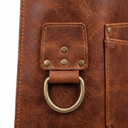 "Extreme close-up of brown leather work apron detail, showing a brass D-ring attached to a stitched leather tab with rivets, along with visible stitching and pocket edge on textured distressed leather."