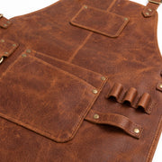 "Close-up detail shot of brown distressed leather work apron showing pocket stitching, brass rivets, front pocket with pen slot, and a multi-loop tool holder for pens or brushes."