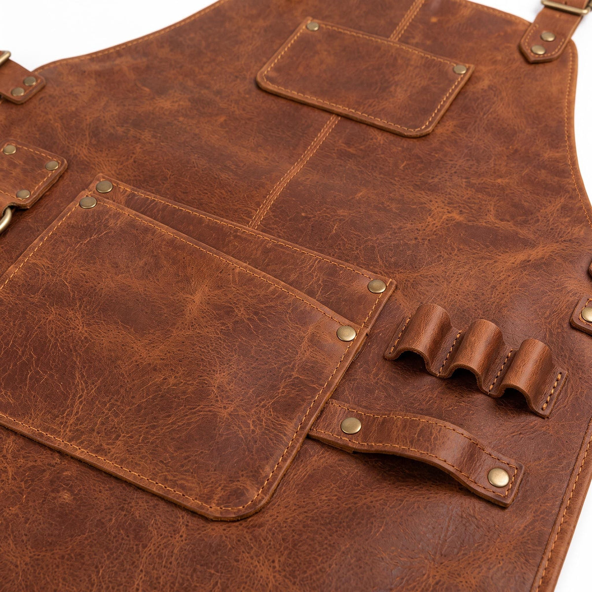 "Close-up detail shot of brown distressed leather work apron showing pocket stitching, brass rivets, front pocket with pen slot, and a multi-loop tool holder for pens or brushes."