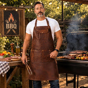 Man wearing brown leather BBQ apron with pockets holding grilling tongs at outdoor barbecue grill