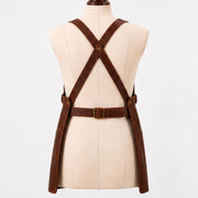 Back view of a brown leather work apron on a mannequin, showing crossback X-strap design with a brass rivet at the center cross point, an adjustable brass buckle waist belt, and two long apron panels hanging down."