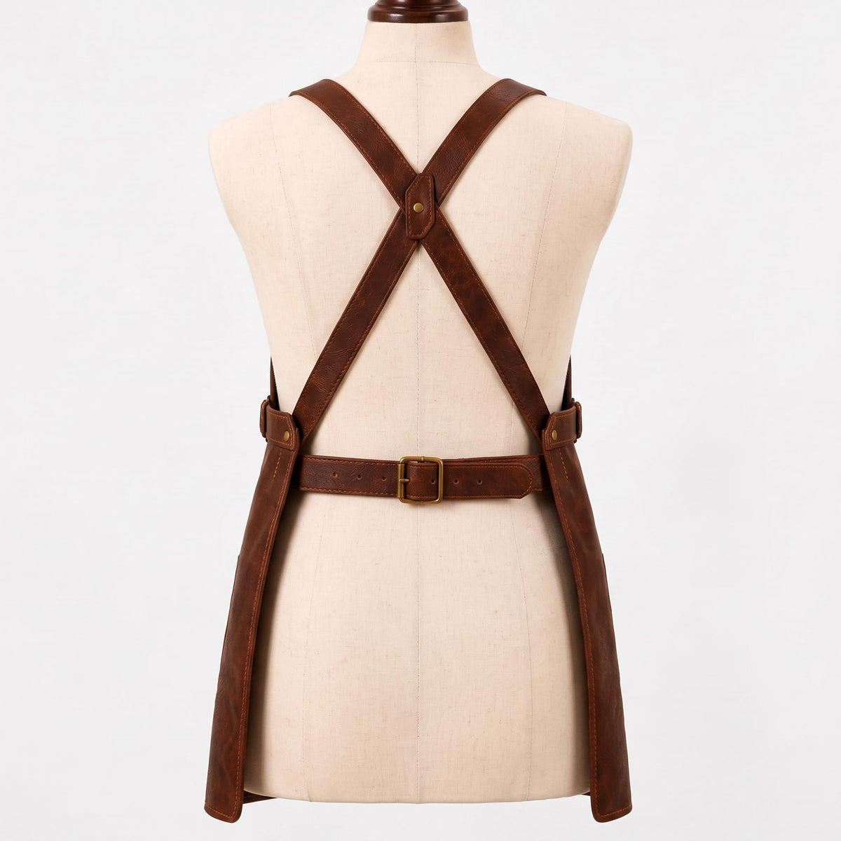 Back view of a brown leather work apron on a mannequin, showing crossback X-strap design with a brass rivet at the center cross point, an adjustable brass buckle waist belt, and two long apron panels hanging down."
