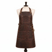 "Dark brown leather apron on a cream-colored mannequin torso, featuring an adjustable neck strap with brass buckle and rivets, a cross-back waist strap with a brass D-ring, a single chest pocket, and a wide pocket panel with multiple compartments at the front lower section, set against a plain white background.