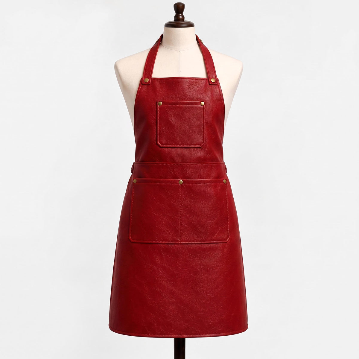 "Red leather bib apron displayed on a cream-colored dress form mannequin against a white background. The apron features a halter neck strap with brass rivets, a square chest pocket, and a wide waist pocket with a center divider, all secured with brass snap buttons. The leather has a rich burgundy-red tone with visible natural grain texture."