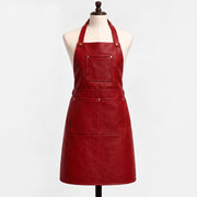 "Red leather bib apron displayed on a cream-colored dress form mannequin against a white background. The apron features a halter neck strap with brass rivets, a square chest pocket, and a wide waist pocket with a center divider, all secured with brass snap buttons. The leather has a rich burgundy-red tone with visible natural grain texture."