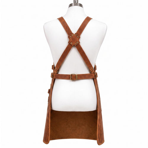 "Back view of brown leather work apron on mannequin, showing crossback straps with X-shaped brass-riveted leather patch, adjustable buckle waist strap, and side buckle straps, against a white background."