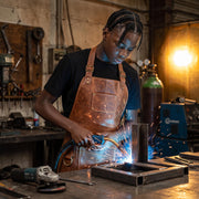 "Young welder wearing brown leather work apron over black t-shirt, using a welding torch on a metal frame in a workshop, with sparks flying and tools, a grinder, and gas cylinders visible in the background."