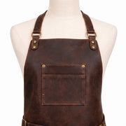 "Close-up detail of a dark brown leather apron's neckline on a cream mannequin, showing adjustable neck straps with brass D-rings and rivets, and a chest pocket with brass rivets and contrast tan stitching, against a plain white background."