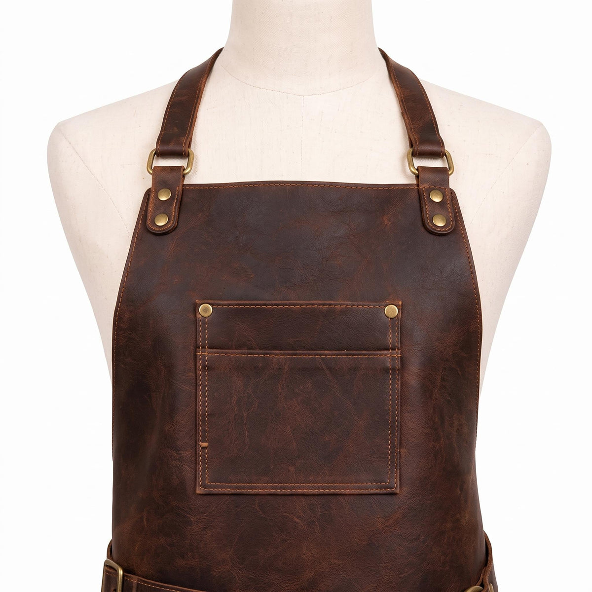 "Close-up detail of a dark brown leather apron's neckline on a cream mannequin, showing adjustable neck straps with brass D-rings and rivets, and a chest pocket with brass rivets and contrast tan stitching, against a plain white background."