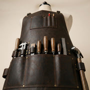 Close-up view of leather woodworking apron tool storage with wood-handled chisels, ruler, caliper and hammer held in pockets and loops.
