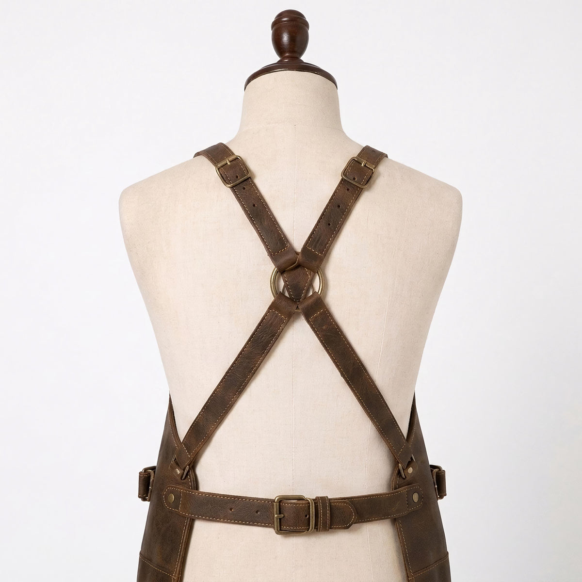 "Close-up of distressed brown leather apron's crisscross X-back straps with brass buckles, brass O-ring, and adjustable waist belt, shown on a mannequin torso."
