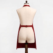 "Back view of a red leather bib apron on a cream-colored dress form mannequin against a white background. Shows an adjustable neck strap with a brass buckle and hole punches, an adjustable waist belt with a brass buckle at the center back, and open sides with the apron falling into two long panels below the waist."