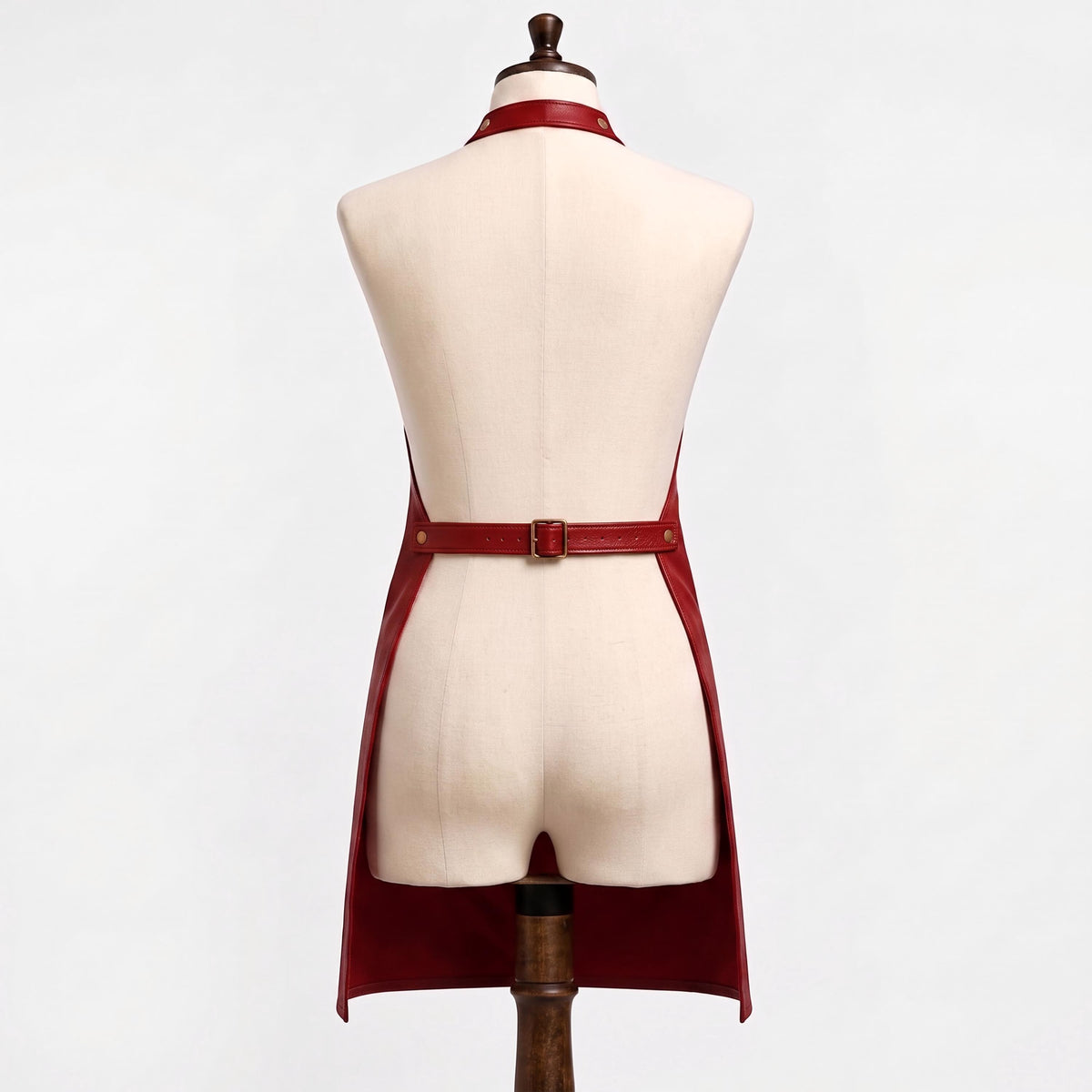 "Back view of a red leather bib apron on a cream-colored dress form mannequin against a white background. Shows an adjustable neck strap with a brass buckle and hole punches, an adjustable waist belt with a brass buckle at the center back, and open sides with the apron falling into two long panels below the waist."