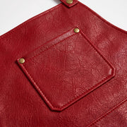 "Extreme close-up of red leather apron pocket detail, showing a diamond-shaped stitched pocket flap secured with two brass snap buttons, along with a neck strap tab fastened with a brass rivet at the top. The leather surface displays detailed natural grain and texture."