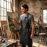"Artist wearing black leather apron with brush pockets while painting on canvas in a sunlit studio."