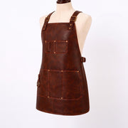 "Three-quarter angle view of a brown leather work apron on a mannequin, showing adjustable brass buckle shoulder straps, a chest pocket with pen slots, side waist buckle, a brass D-ring, and a large stitched front pocket."