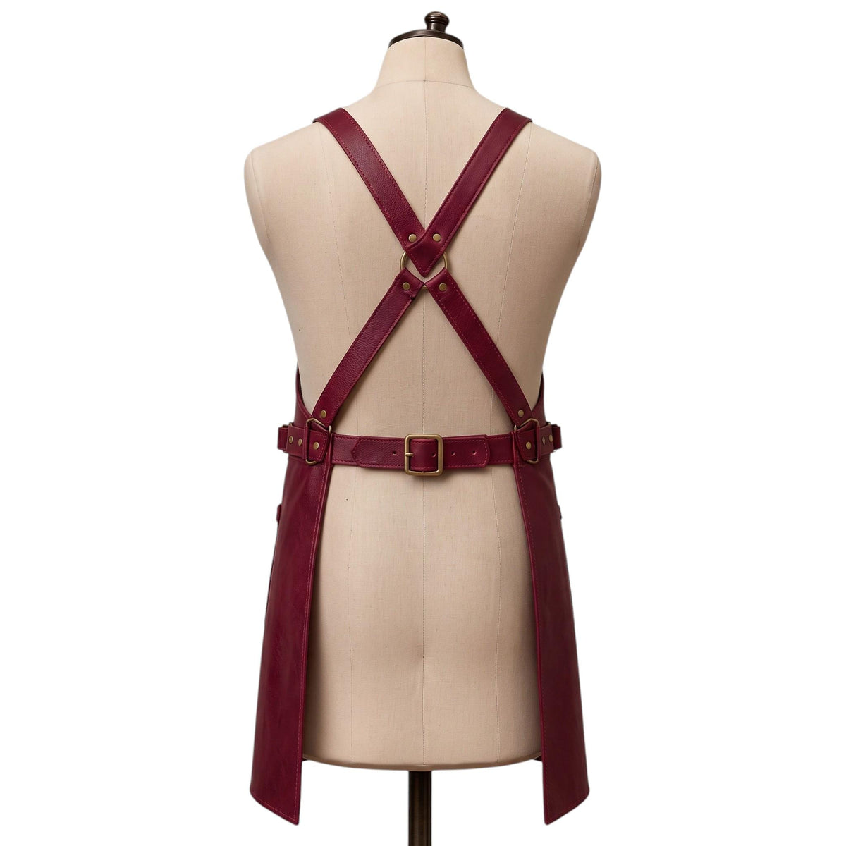 "Back view of a burgundy leather apron with crossback straps, a brass D-ring, and an adjustable waist buckle."