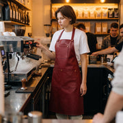 "A young barista with short brown hair, wearing a red leather bib apron with a chest pocket and wide front pocket over a white t-shirt, adjusts the steam wand knob on a espresso machine in a busy coffee shop. Shelves stocked with coffee bean bags line the wall in the warmly lit background, with other people visible at the counter."