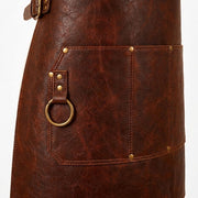 "Close-up detail of a brown leather apron's front pocket, showing riveted stitching, a leather loop with an antique brass D-ring for hanging tools, and a side buckle strap visible at the top."