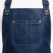 "Close-up of navy blue leather apron's top section, showing brass buckle straps with rivets, contrast stitching, and a chest pocket with visible topstitch detailing and brass snap closures."