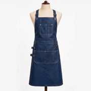 "Navy blue leather apron displayed on a fabric mannequin against a white background, featuring adjustable brass buckle shoulder straps, a large chest pocket with contrast stitching, a waist belt with a brass D-ring, and a lower double pocket with a small side loop for tools."