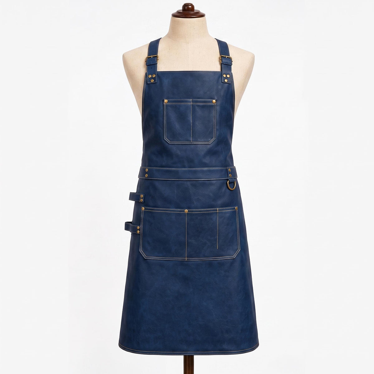 "Navy blue leather apron displayed on a fabric mannequin against a white background, featuring adjustable brass buckle shoulder straps, a large chest pocket with contrast stitching, a waist belt with a brass D-ring, and a lower double pocket with a small side loop for tools."
