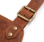 "Extreme close-up of brown leather work apron's brass roller buckle with adjustment holes, and a riveted leather tab with two brass rivets, showing detailed stitching on textured leather."