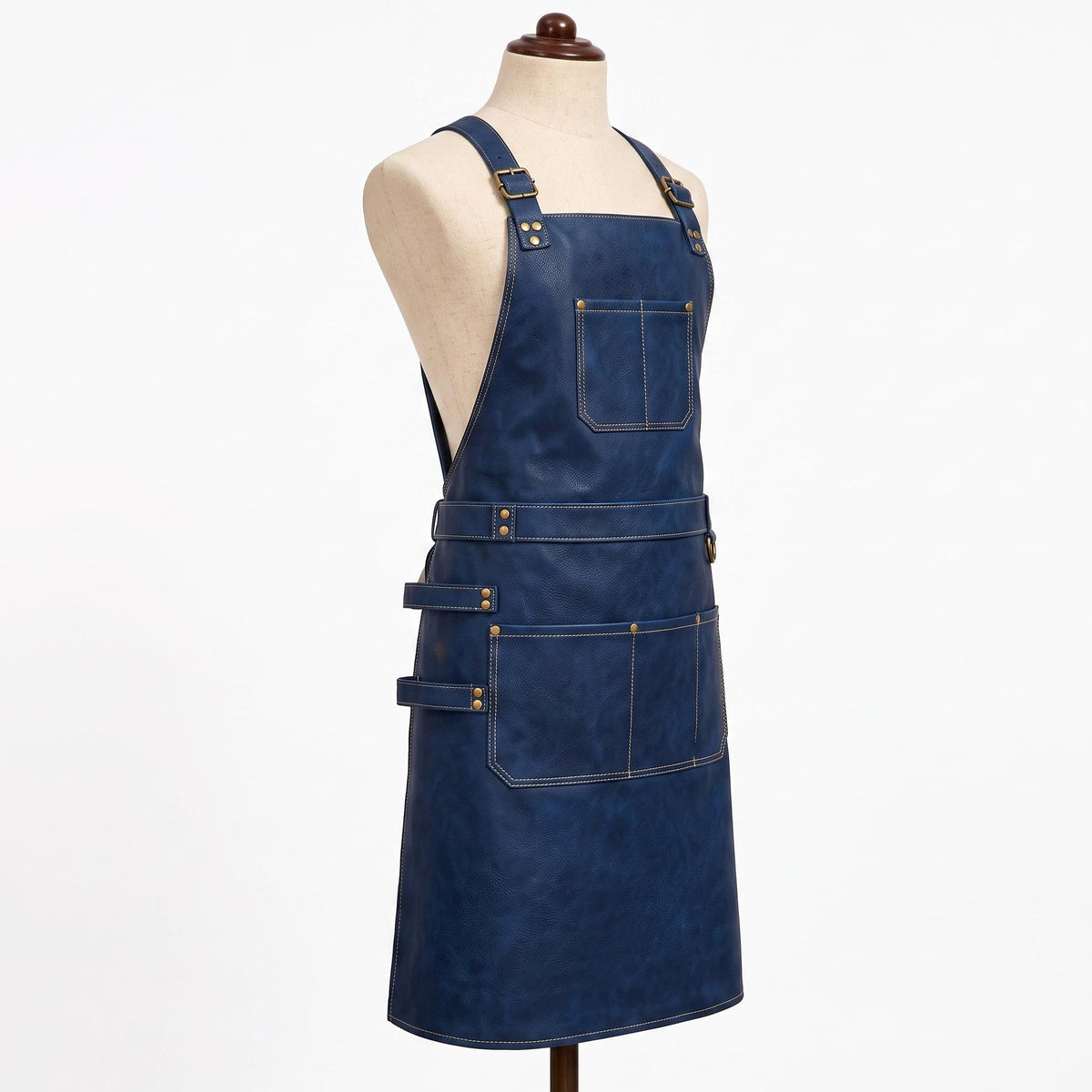 side angle view): "Side-angled view of navy blue leather apron on a fabric mannequin, showing adjustable brass buckle straps, a chest pocket, waist belt with D-ring, and lower pocket with a side loop for tools