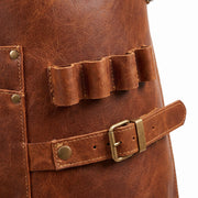 "Close-up detail of brown leather work apron showing a multi-loop tool holder for pens or utensils, brass roller buckle waist strap with adjustment holes, and riveted stitched panels on textured leather."