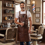 Smiling barber wearing a brown leather tool apron in a vintage-style barbershop."