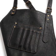 "Close-up of black leather apron showing chest pocket stitching and brass buckle strap detail."