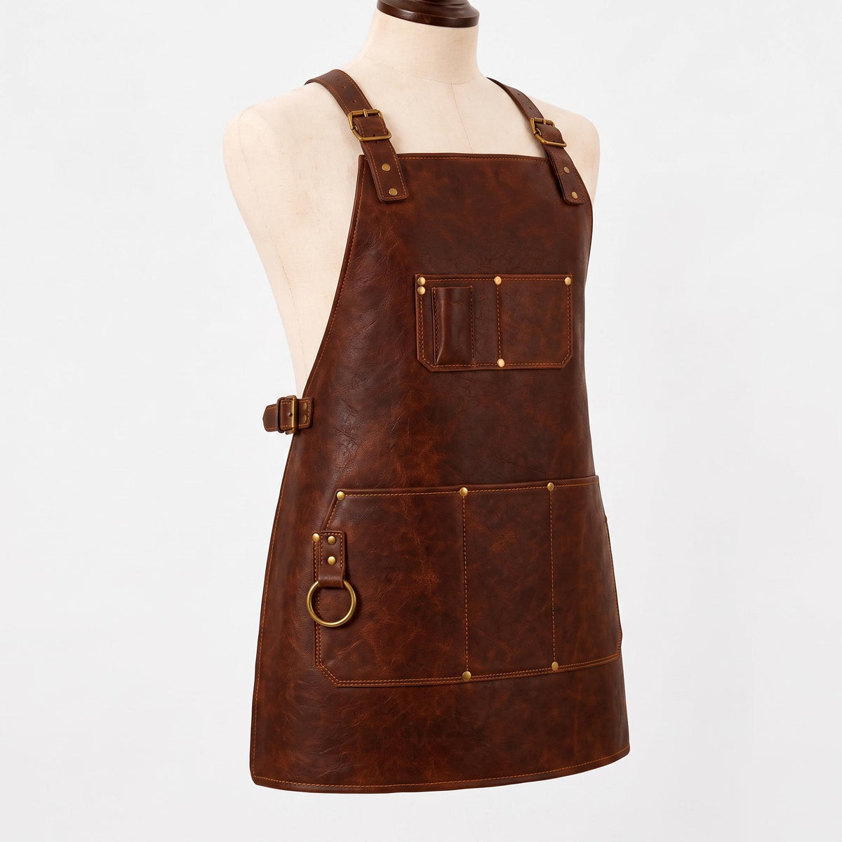 "Angled side view of a brown leather work apron on a mannequin, showing adjustable brass buckle straps, a chest pocket with pen slots, side waist buckle, a brass D-ring for tools, and a large front pocket with stitched compartments."