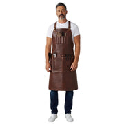A man wearing a dark brown leather apron with multiple pockets and tool loops over a white t-shirt, blue jeans, and white sneakers, standing against a solid white background.