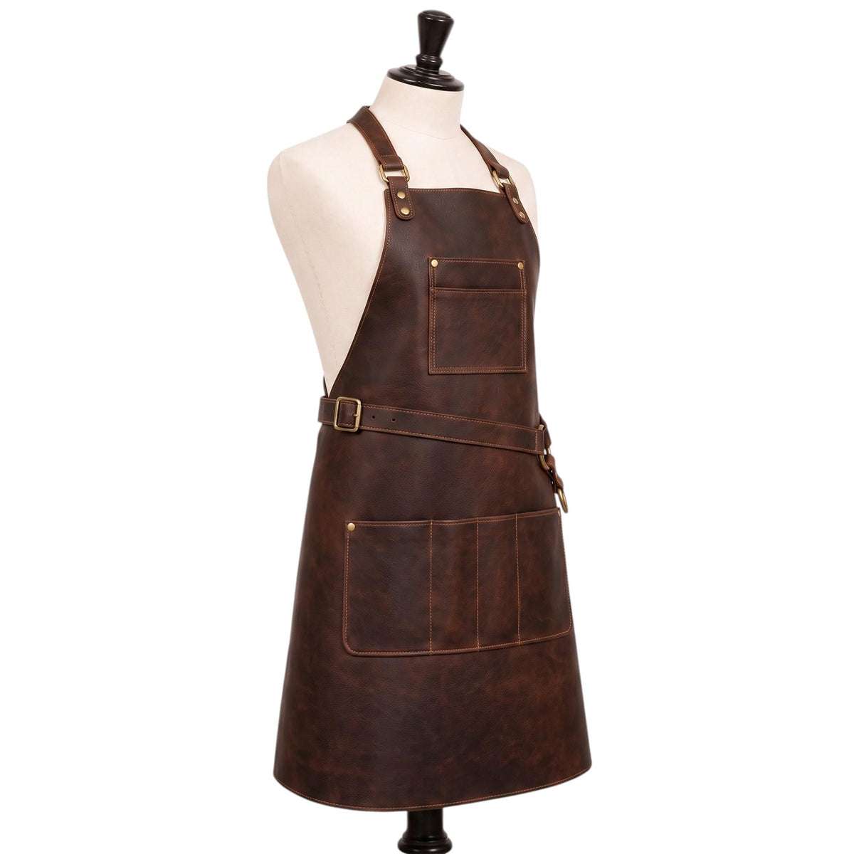 "Side-angle view of a dark brown leather apron on a cream mannequin torso, showing the adjustable neck strap with brass buckle, a diagonal cross-back waist strap secured with a brass ring, a chest pocket, and a multi-compartment lower pocket, against a plain white background."