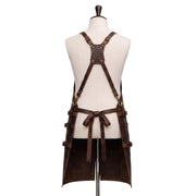 "Back view of a dark brown leather apron displayed on a fabric mannequin, showing an X-shaped cross-back strap design with brass buckles, a central reinforced leather patch, adjustable side straps with buckles, and a waist tie knotted in a bow, with the apron split into two hanging leg panels at the bottom, photographed against a plain white background."