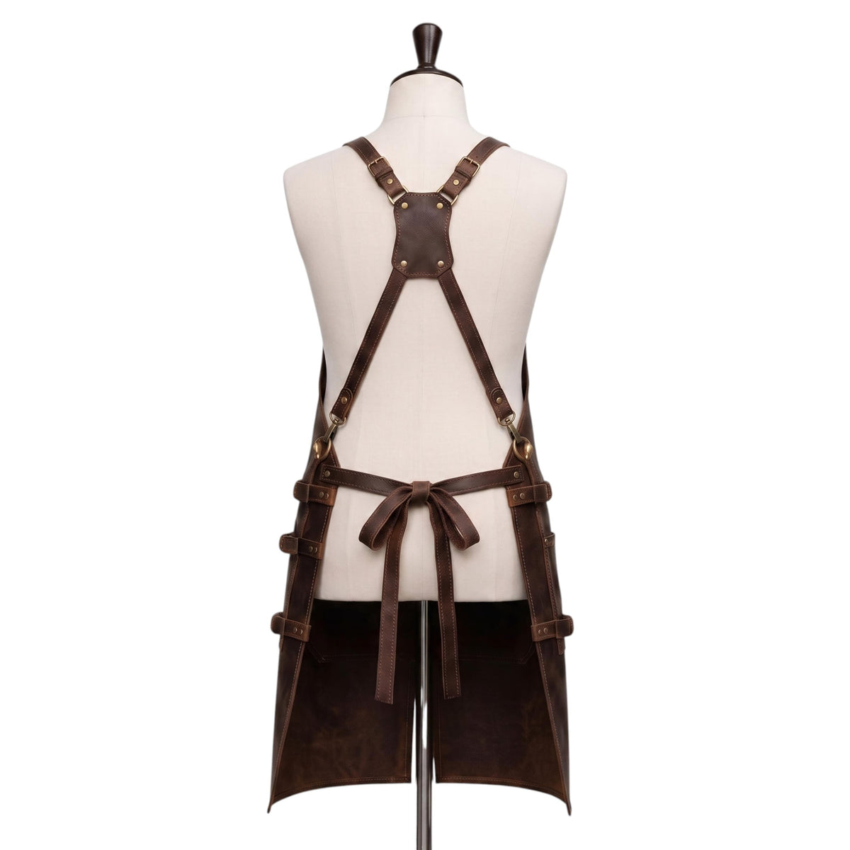 "Back view of a dark brown leather apron displayed on a fabric mannequin, showing an X-shaped cross-back strap design with brass buckles, a central reinforced leather patch, adjustable side straps with buckles, and a waist tie knotted in a bow, with the apron split into two hanging leg panels at the bottom, photographed against a plain white background."