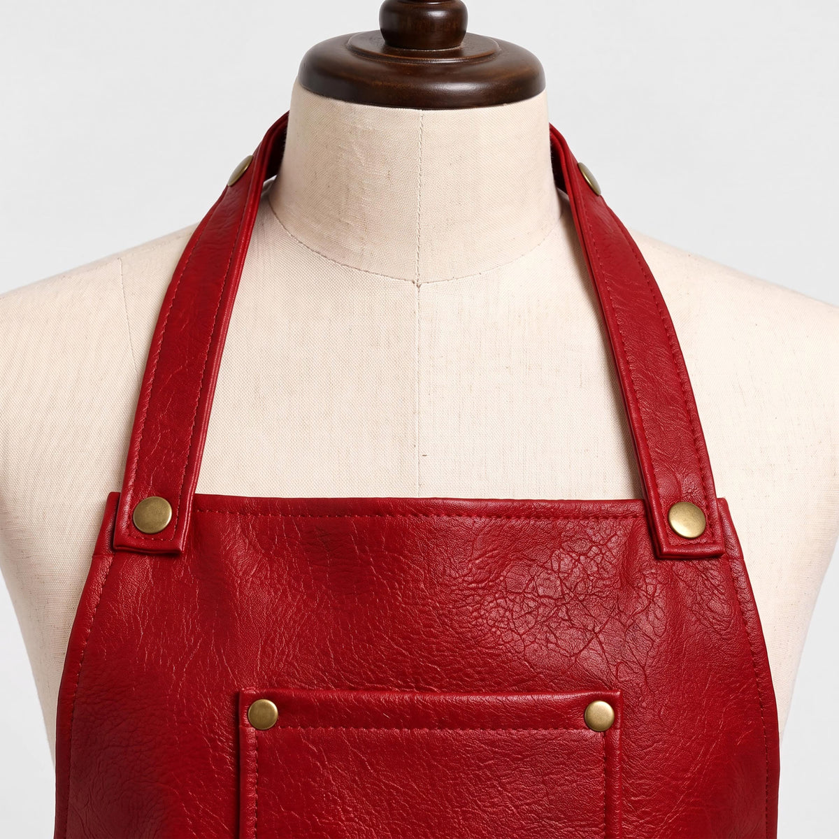 "Close-up view of a red leather bib apron's neck strap and chest area on a cream-colored dress form mannequin. Shows the halter straps attached with brass rivets at the shoulder, visible natural leather grain texture on the bib, and the top corner of a chest pocket with brass snap buttons."