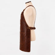 "Side profile view of a brown leather work apron on a mannequin, showing the neck strap buckle, side pocket panel, adjustable waist buckle strap, and a lower front pocket with stitched edges."