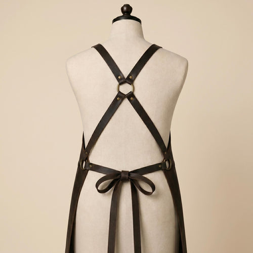 Back view of brown leather work apron showing adjustable cross-back shoulder straps, central metal ring and leather waist tie.