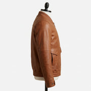 "Full profile side view of a tan brown leather bomber jacket with ribbed collar, flap pocket, and quilted elbow panel, displayed on a mannequin against a light gray background."