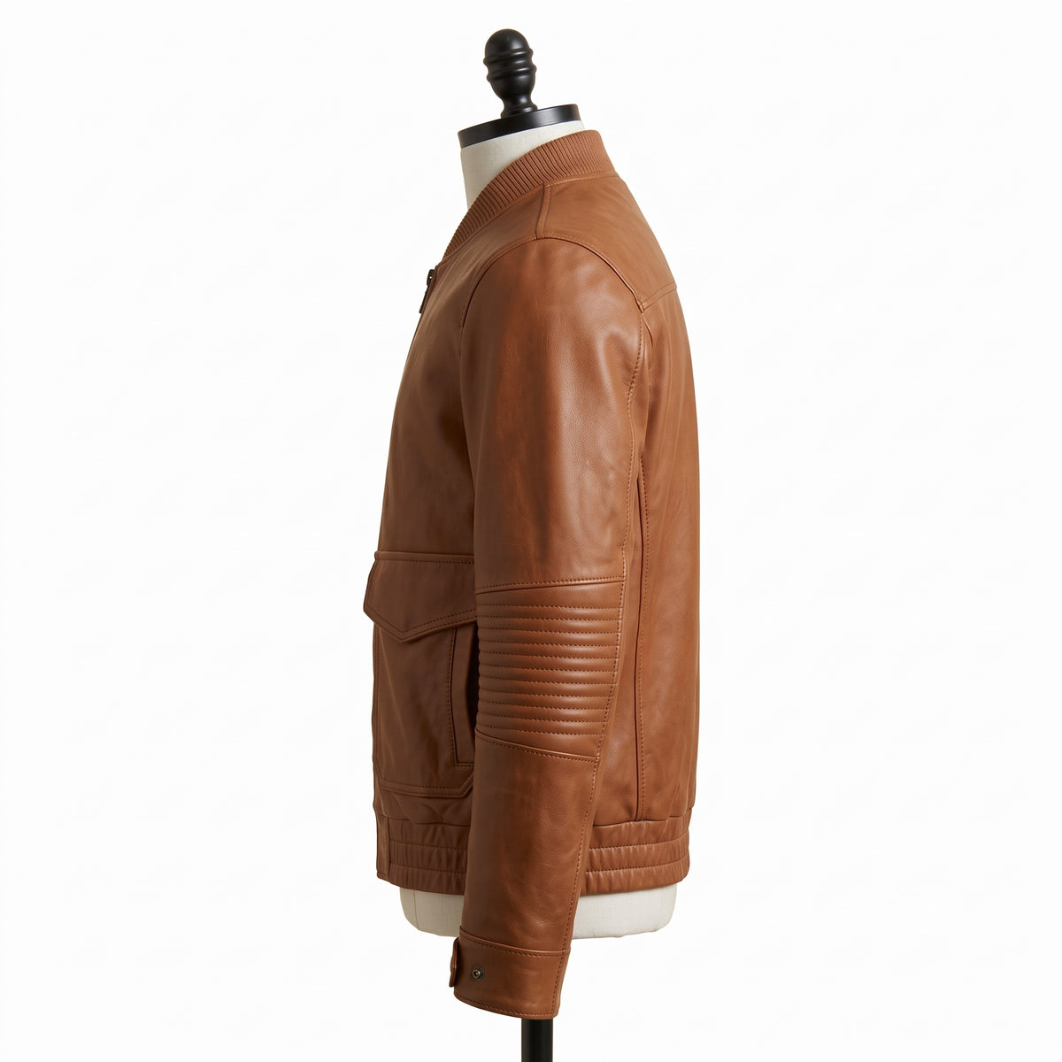 "Full profile side view of a tan brown leather bomber jacket with ribbed collar, flap pocket, and quilted elbow panel, displayed on a mannequin against a white background."