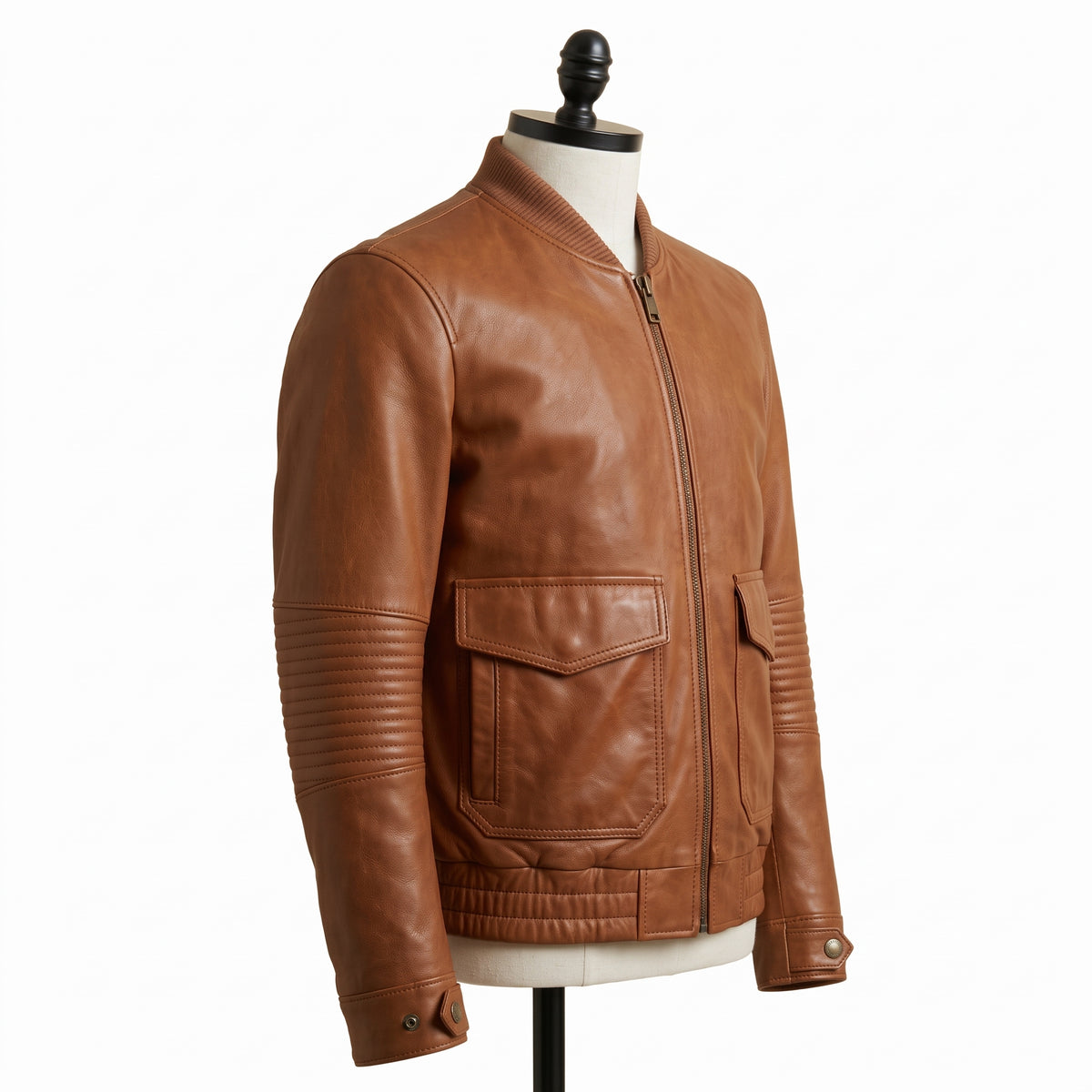 "Angled side view of a tan brown leather bomber jacket with ribbed collar, front zipper, flap pockets, and quilted elbow panels, displayed on a mannequin against a white background."