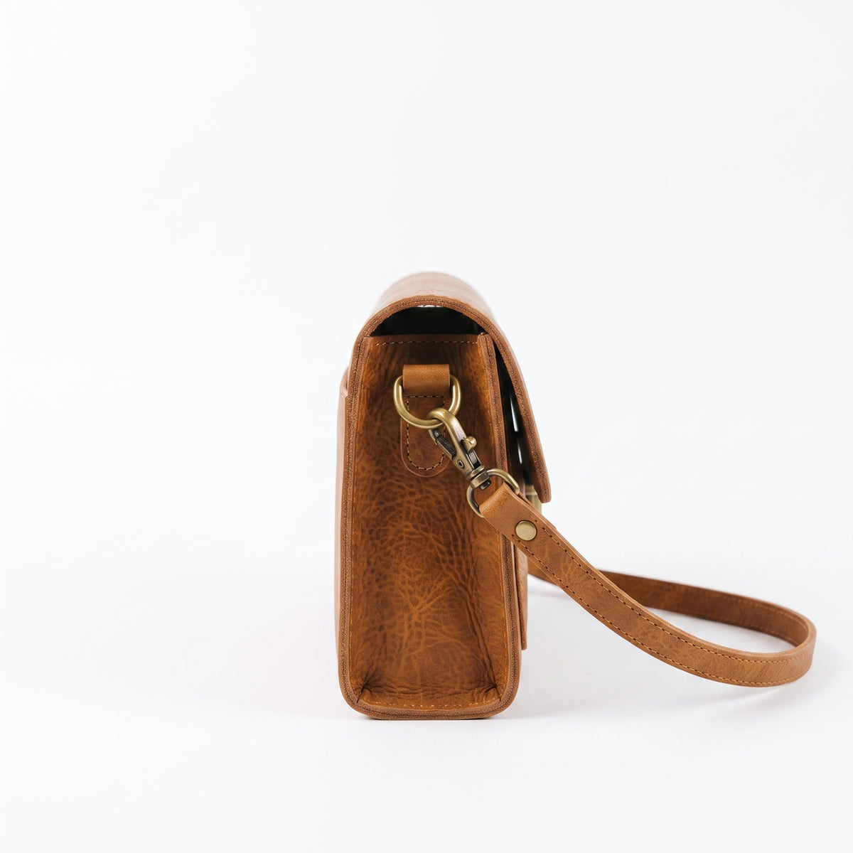 "Side view of a tan leather crossbody bag showing its depth, an open compartment, a gold D-ring and swivel clasp, and the adjustable shoulder strap curving down against a light background."