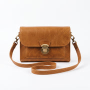 "Front view of a tan leather crossbody bag with a vintage brass push-lock clasp on the flap, and an adjustable strap looped loosely in front against a light background."