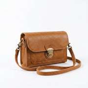 "Angled three-quarter view of a tan leather crossbody bag showing the brass push-lock clasp, adjustable strap with swivel hooks, and leather grain texture against a light background."