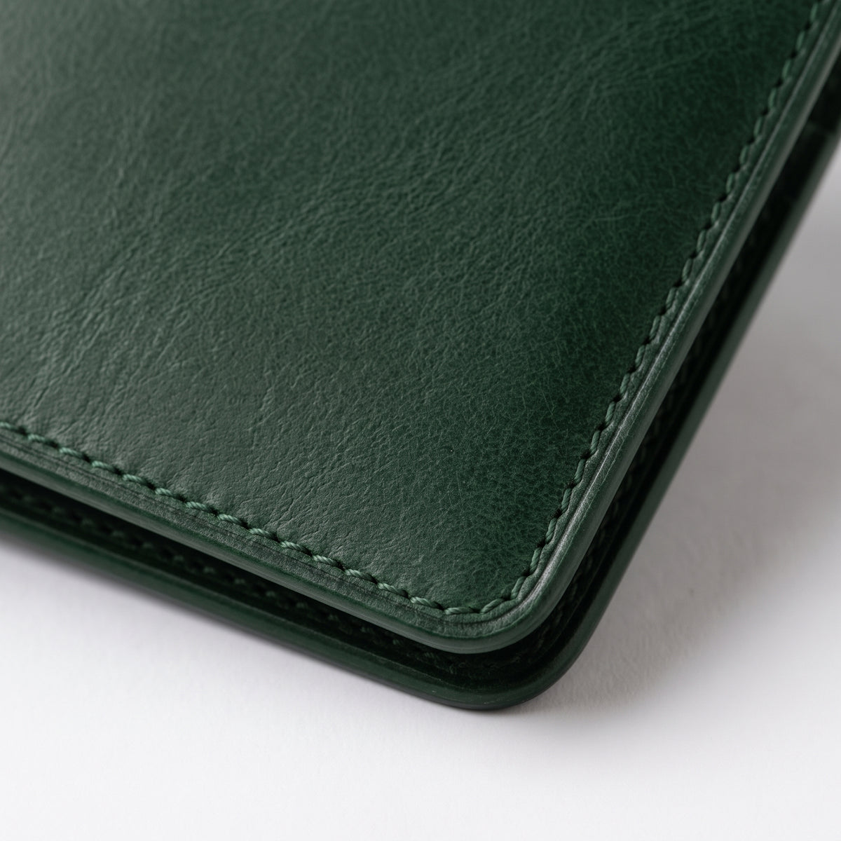Close-up of a dark green textured leather folio or wallet corner with visible stitching and a dark green protective edge, photographed on a white background.