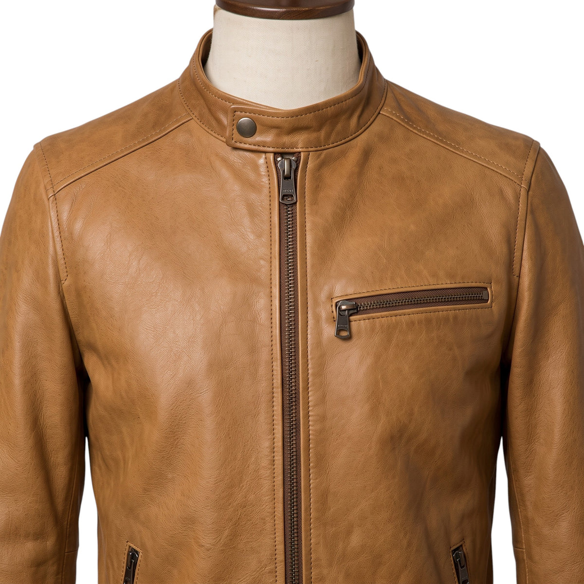 "Close-up of a tan leather biker jacket's collar and chest area on a mannequin, showing the snap button collar, front zipper, and chest zip pocket in detail."