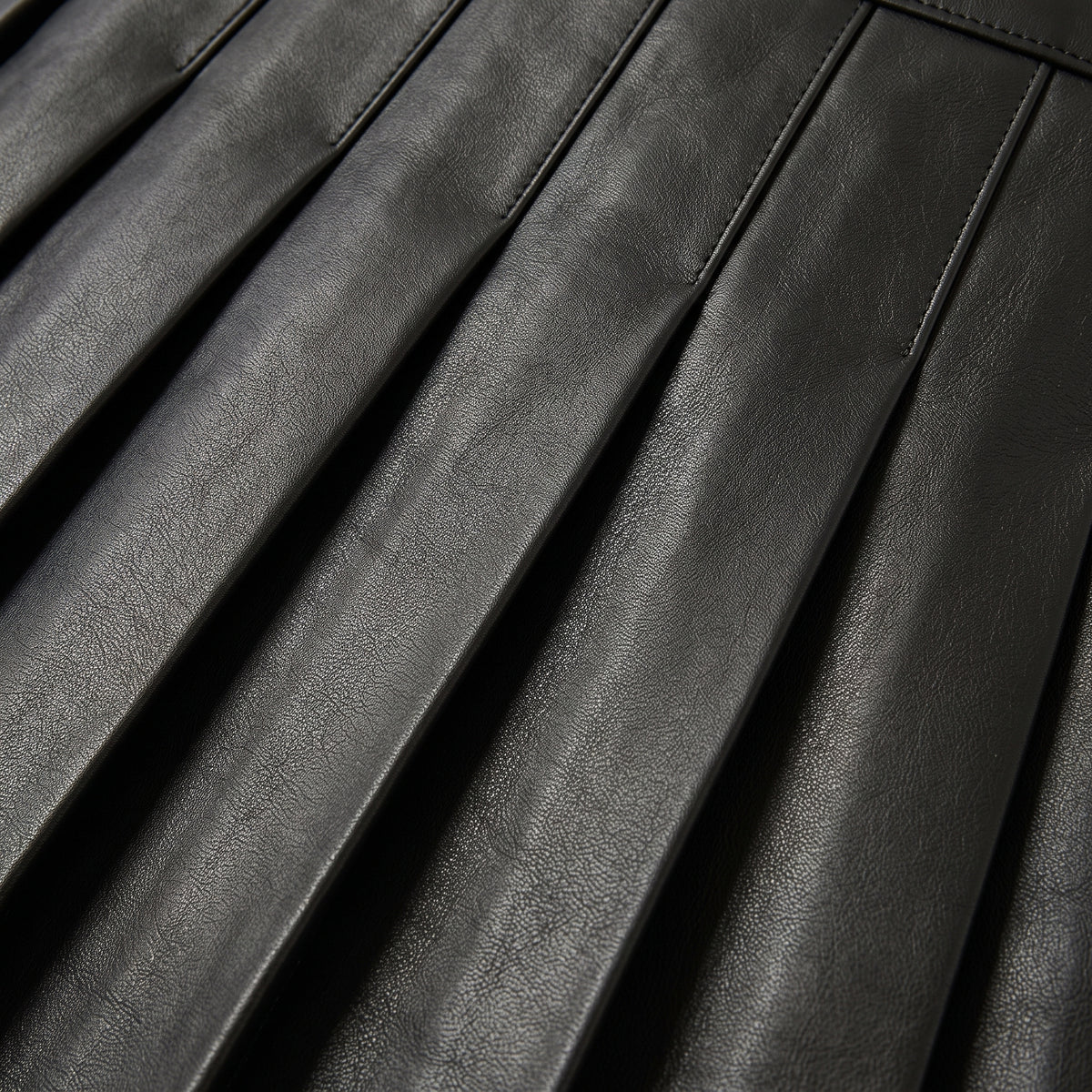 "Extreme close-up of the pleats on a black leather skirt, shot at an angle showing the leather grain texture and stitched pleat lines."