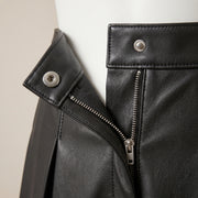 "Extreme close-up of the pleats on a black leather skirt, shot at an angle showing the leather grain texture and stitched pleat lines."