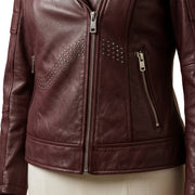 A close-up of a women's burgundy leather jacket with silver zippers and studded details, shown on a mannequin torso against a white background.